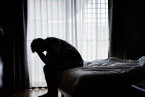 man seated alone on edge of his bed in a darkened room with his head in his hands and wondering what are the risks of tranq