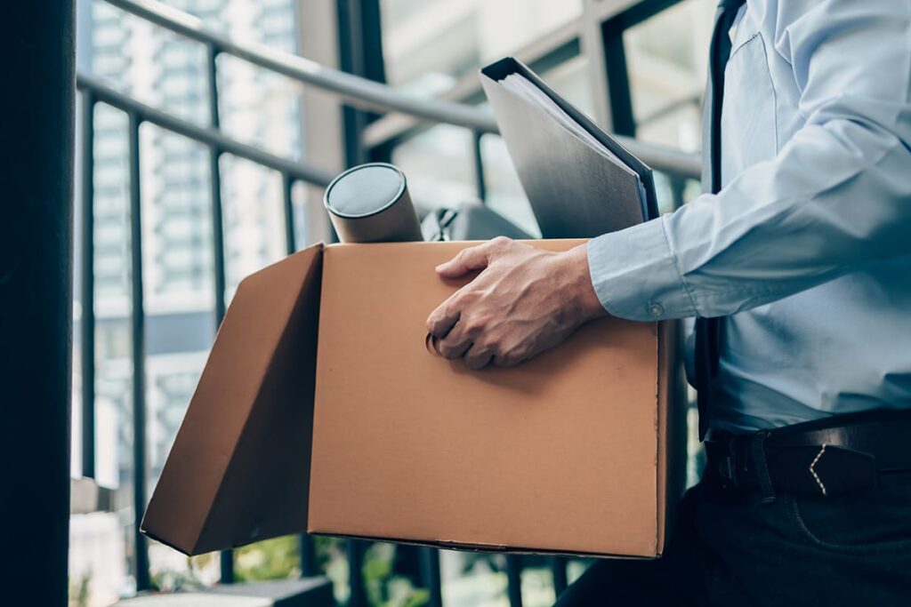 image of businessman carrying box through office after clearing out his desk and discovering how unemployment and mental health are related.