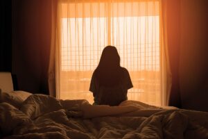 silhouette of young woman seated on her bed in a darkened room in front of window at sunset reflecting on recognizing heroin dependence in her loved ones