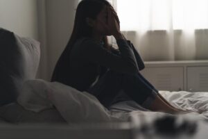 woman seated on her bed in a darkened room wondering can PTSD impact addiction?