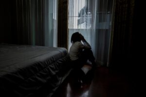 woman seated on her bedroom floor in a darkened room staring out a window at night experiencing the dangers of Adderall and alcohol.