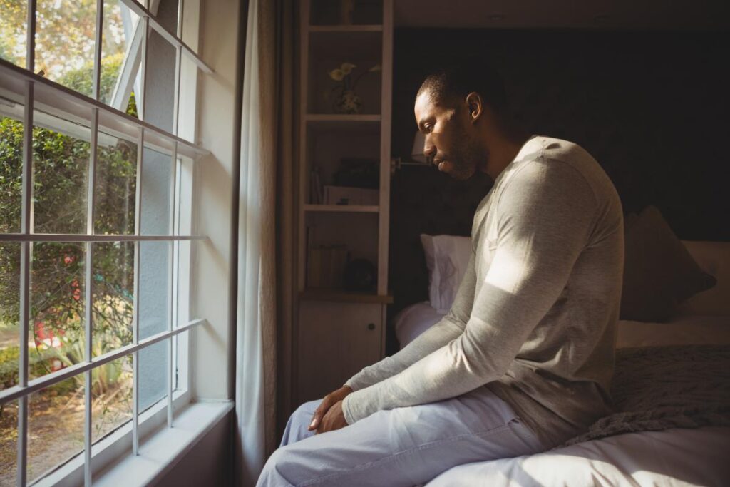 man seated despondently on his bed staring out the widow and suffering the long term effects of heroin.