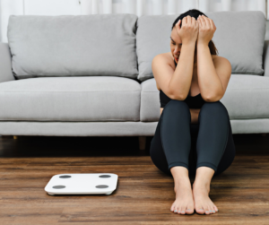 Woman sits on floor next to scale while thinking about the link between alcohol and weight gain